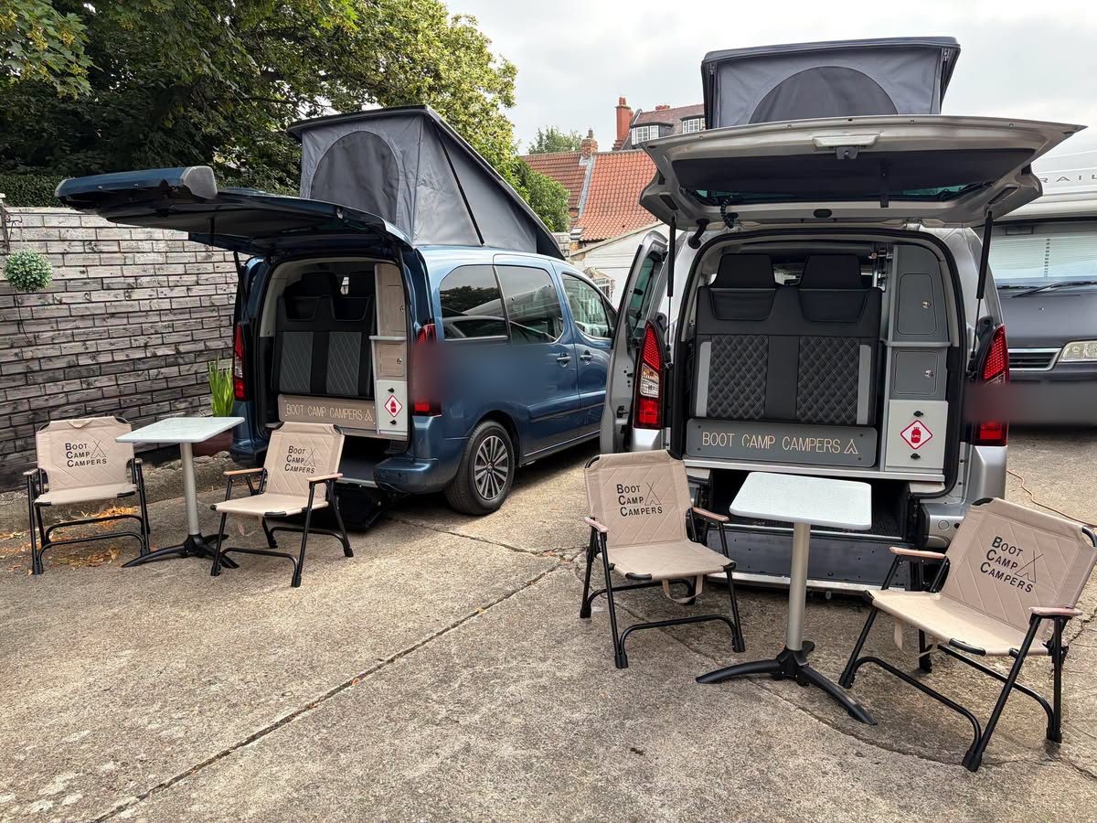 Two Boot Camp Campers vans with pop-top roofs, camping chairs and tables set out on a drive.